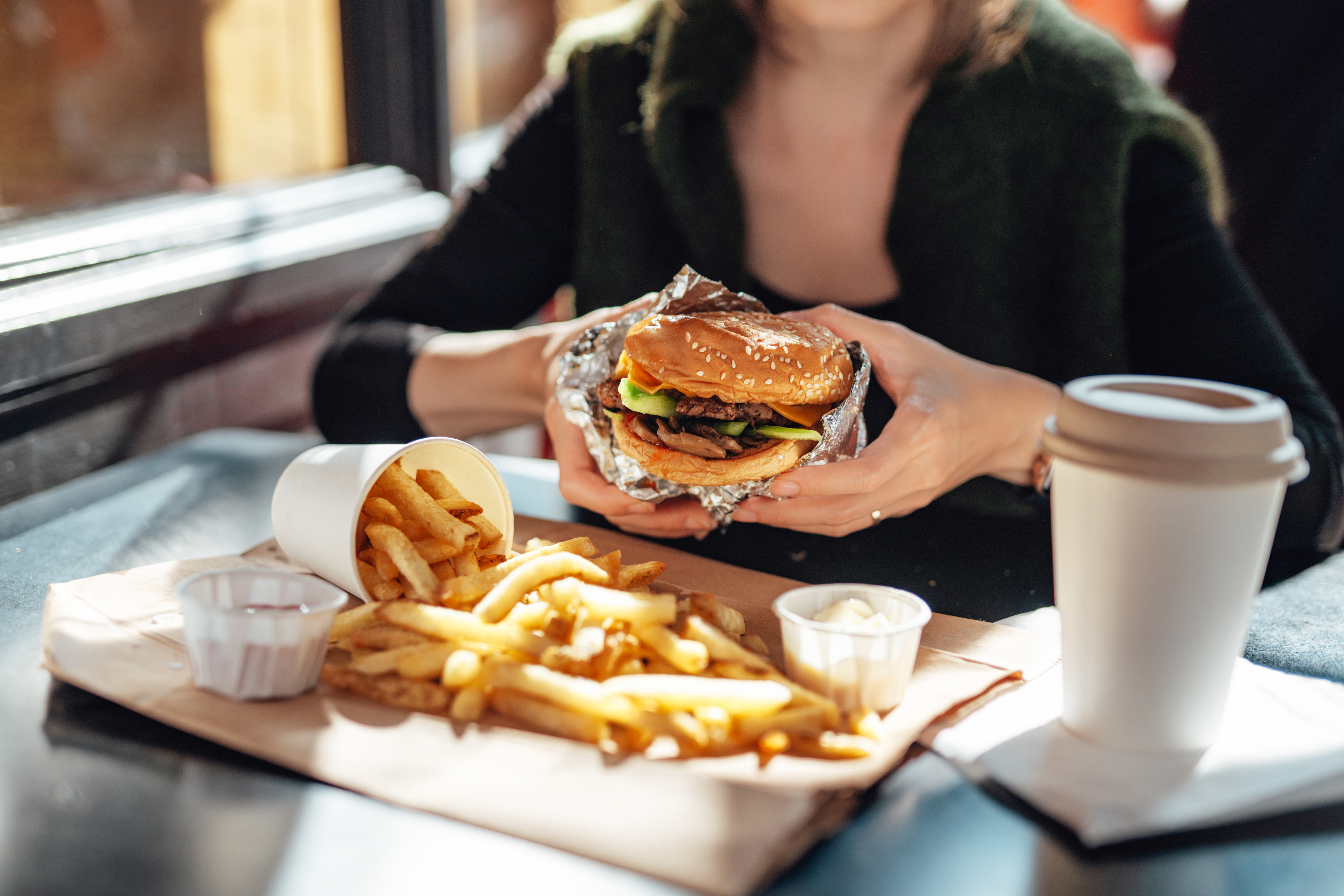 Woman holding large burger in front of a plate of fries and large drink.
